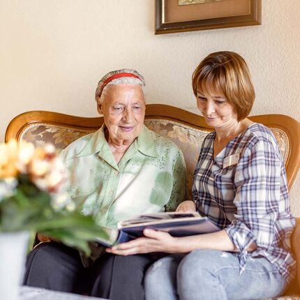 Eine Seniorin und eine Frau sitzen auf einem Sofa und schauen sich ein Fotoalbum an. 