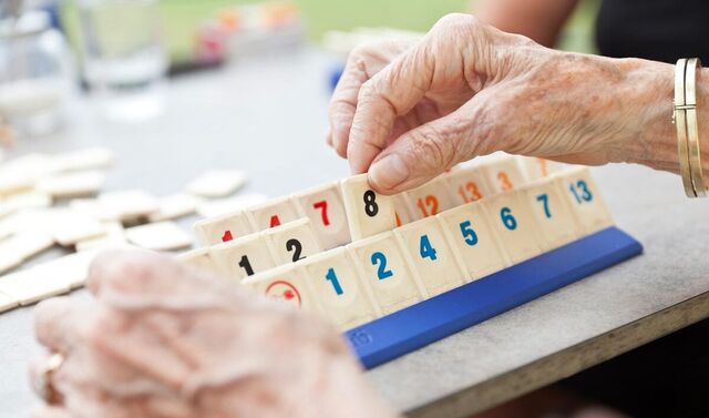 Auf einem Tisch steht ein Rummykub Brettchen mit entsprechenden Spielsteinen darauf. Die Hände einer älteren Frau greifen nach einem Spielstein mit einer Sechs.